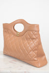 A tan quilted leather handbag with a circular cutout handle and a gold-tone interlocking CC logo, resting on a light marble surface against a white background.