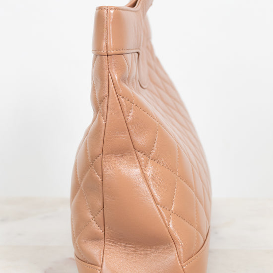 A tan quilted leather shoulder bag shown from a side profile view, resting on a light marble surface against a white background.