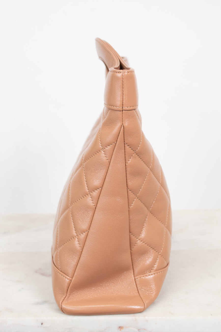 A tan quilted leather shoulder bag shown from a side profile view, resting on a light marble surface against a white background.