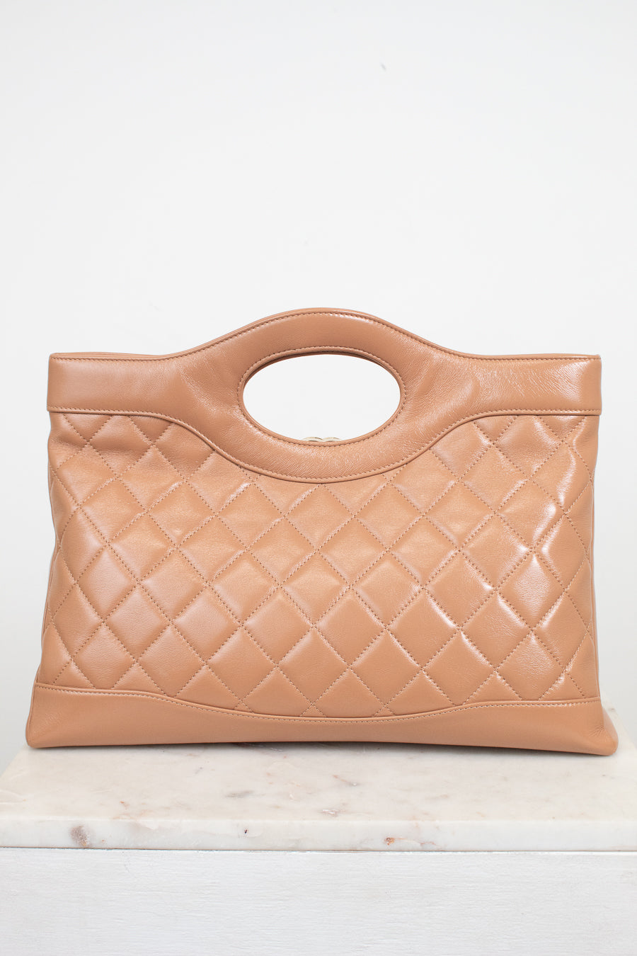 A tan quilted leather handbag with a top handle, displayed on a light marble surface against a white background.