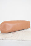 A tan lambskin leather clutch bag with a rounded rectangular shape, resting on a white marble surface against a white background.