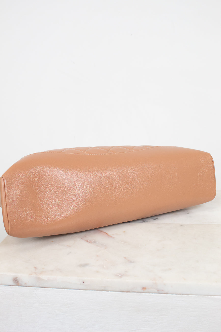 A tan lambskin leather clutch bag with a rounded rectangular shape, resting on a white marble surface against a white background.