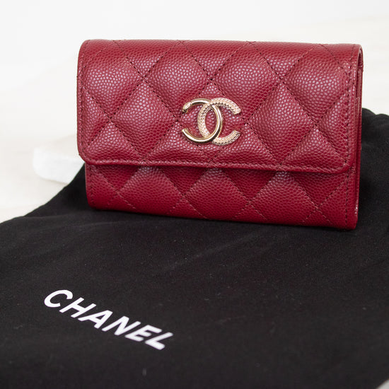 A red Chanel wallet with a quilted caviar texture and a gold-tone interlocking CC logo, resting on a black Chanel dust bag over a light marble surface.