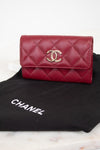 A red Chanel wallet with a quilted caviar texture and a gold-tone interlocking CC logo, resting on a black Chanel dust bag over a light marble surface.