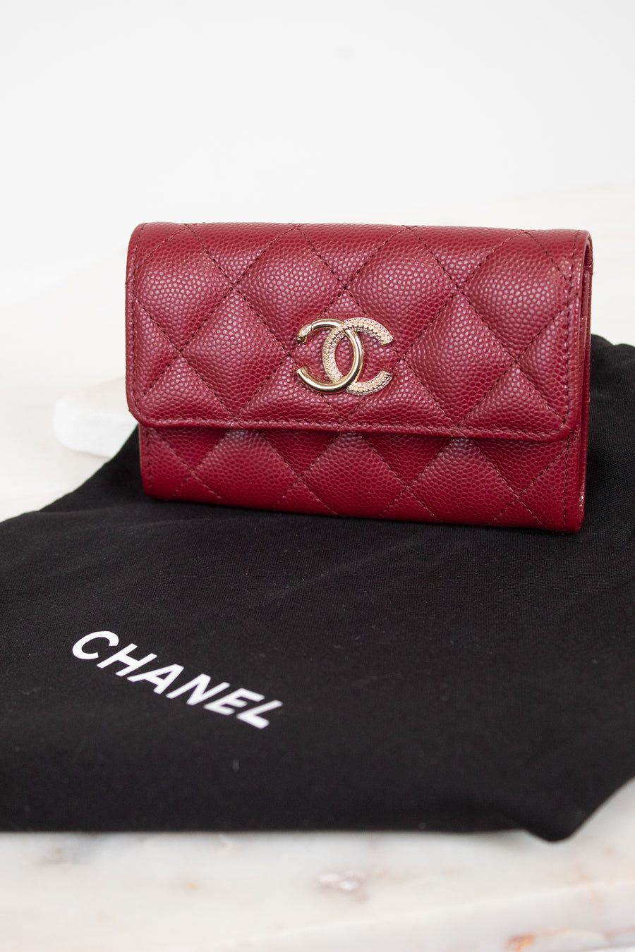 A red Chanel wallet with a quilted caviar texture and a gold-tone interlocking CC logo, resting on a black Chanel dust bag over a light marble surface.