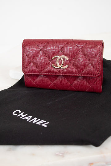A red Chanel wallet with a quilted caviar texture and a gold-tone interlocking CC logo, resting on a black Chanel dust bag over a light marble surface.