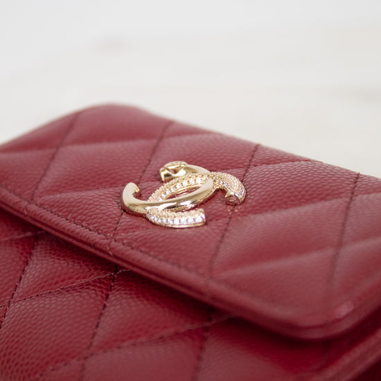 A close-up of a quilted red leather wallet featuring a gold-tone interlocking clasp embellished with small clear crystals, set against a light neutral background.