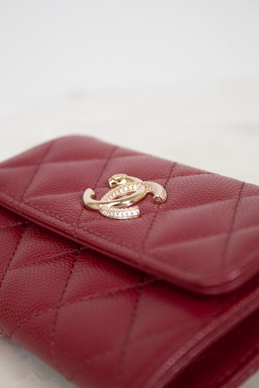 A close-up of a quilted red leather wallet featuring a gold-tone interlocking clasp embellished with small clear crystals, set against a light neutral background.