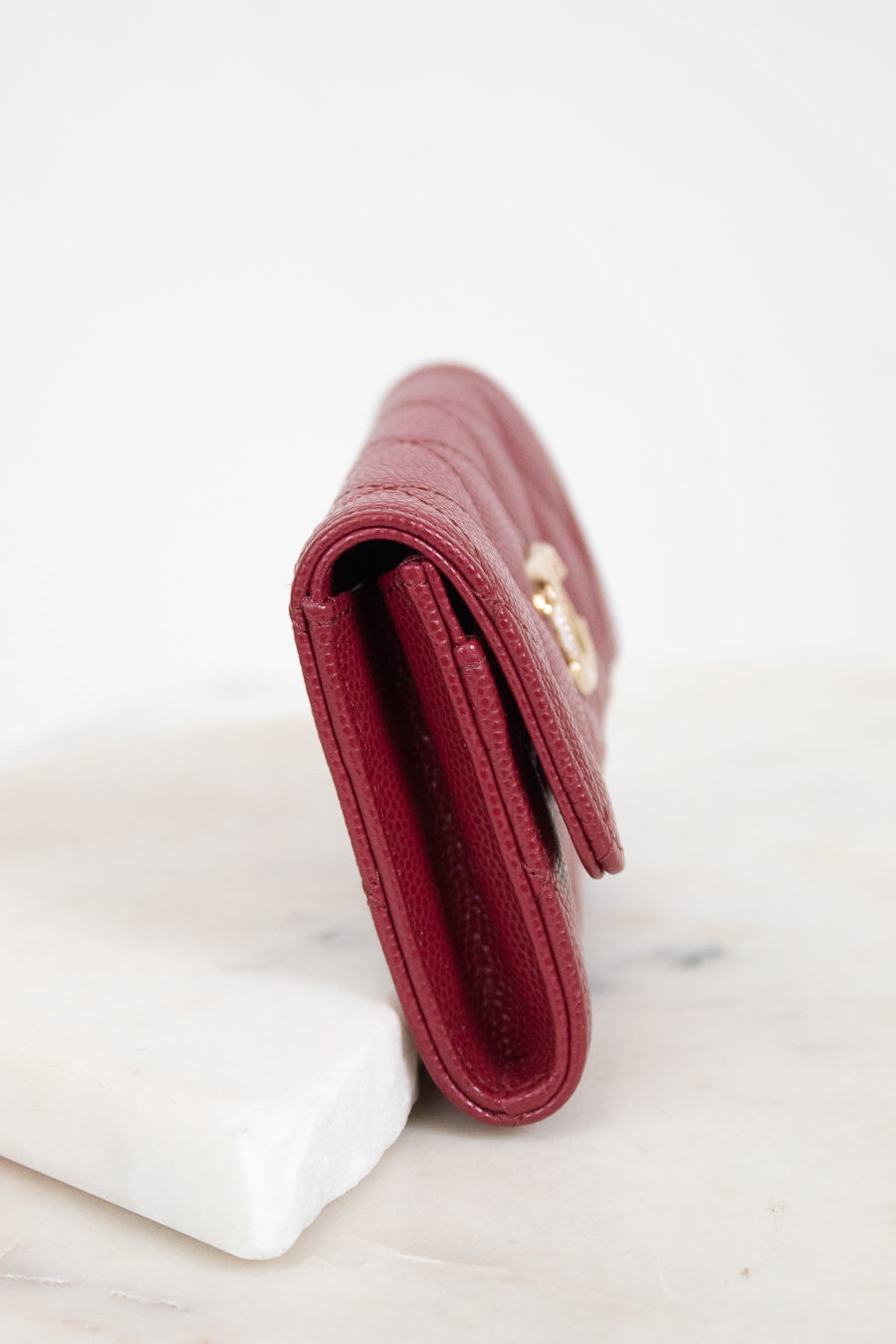 A close-up side view of a burgundy textured leather wallet resting on a white marble block against a white background.