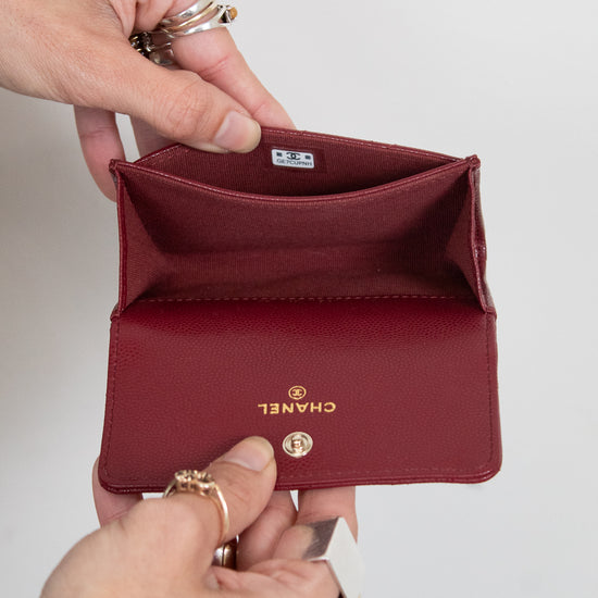 A close-up of a person's hands holding open a small Chanel bifold wallet in deep burgundy leather. The brand name 'CHANEL' is printed in gold on the front flap, and a gold turn-lock clasp is visible.