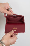 A close-up of a person's hands holding open a small Chanel bifold wallet in deep burgundy leather. The brand name 'CHANEL' is printed in gold on the front flap, and a gold turn-lock clasp is visible.