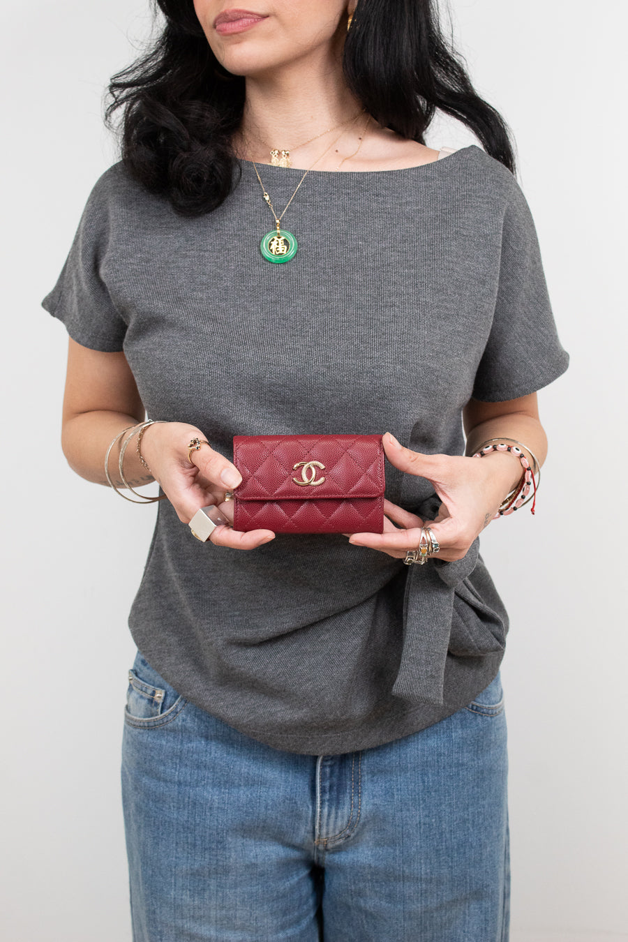 A woman wearing a gray t-shirt and blue jeans holds a small red quilted Chanel wallet with a gold CC logo.