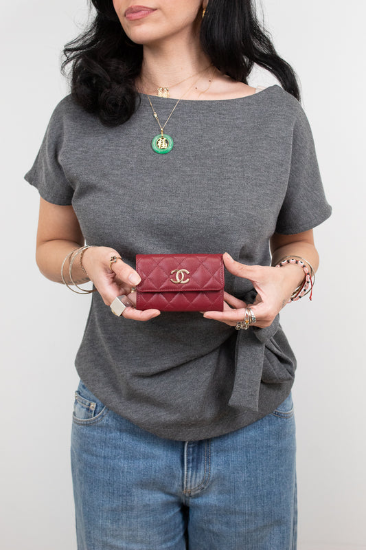 A woman wearing a gray t-shirt and blue jeans holds a small red quilted Chanel wallet with a gold CC logo.