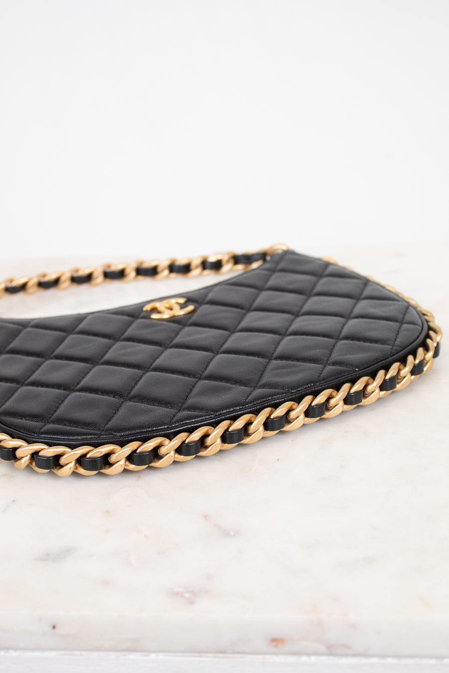 A close-up of a black quilted leather handbag with a gold chain strap and a gold interlocking CC logo, resting on a white marble surface.