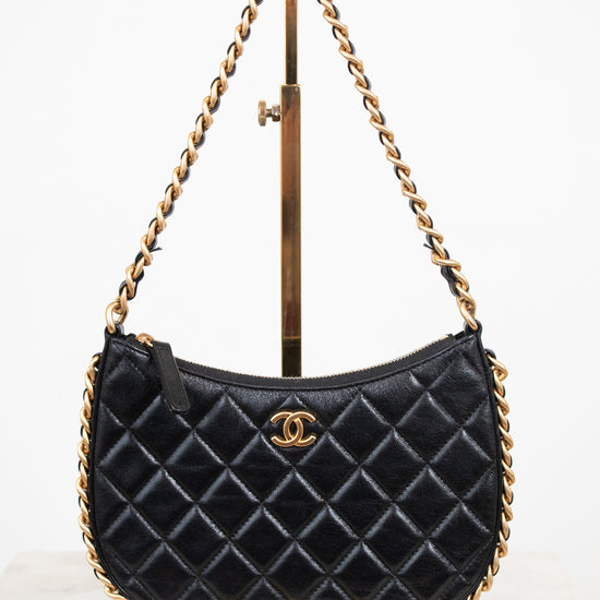 A black quilted leather shoulder bag with a gold-tone chain strap and a gold interlocking CC logo, displayed on a gold stand against a white background.