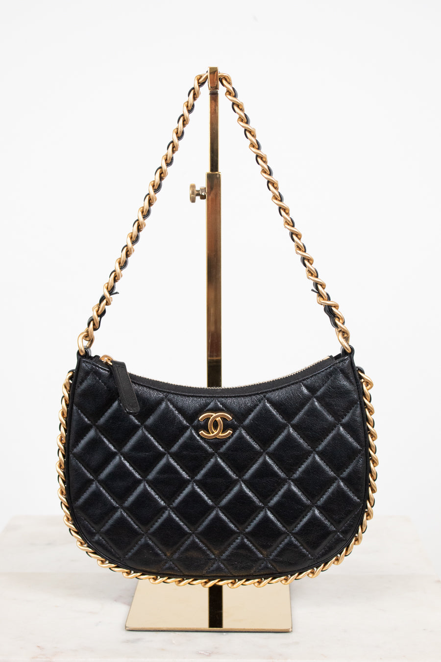 A black quilted leather shoulder bag with a gold-tone chain strap and a gold interlocking CC logo, displayed on a gold stand against a white background.