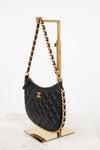 A black quilted leather shoulder bag with gold-tone chain strap and a gold interlocking CC logo, displayed on a gold stand against a white background.