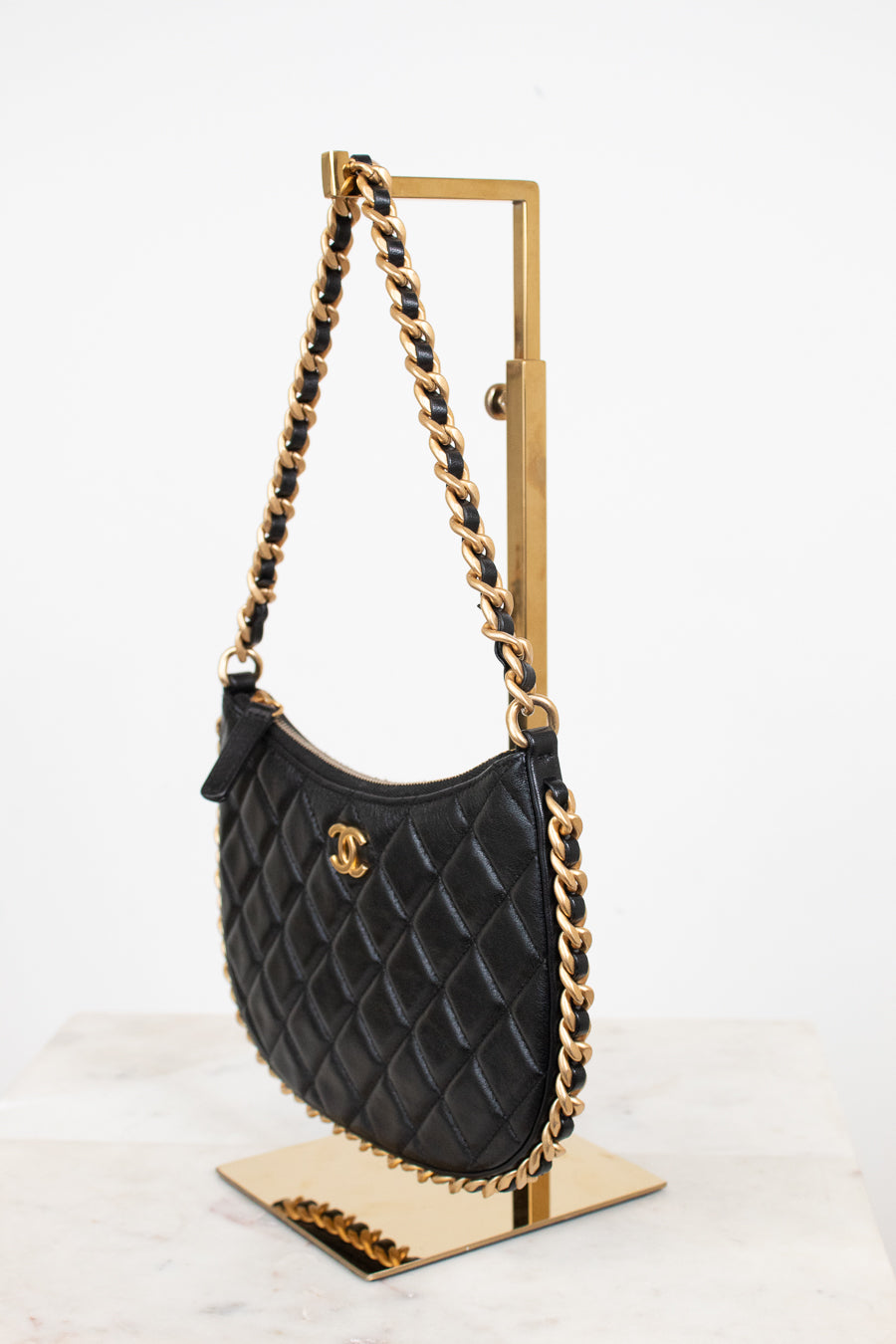 A black quilted leather shoulder bag with gold-tone chain strap and a gold interlocking CC logo, displayed on a gold stand against a white background.