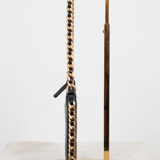 A black leather handbag with a prominent gold chain strap, displayed on a polished gold metal stand against a white background.