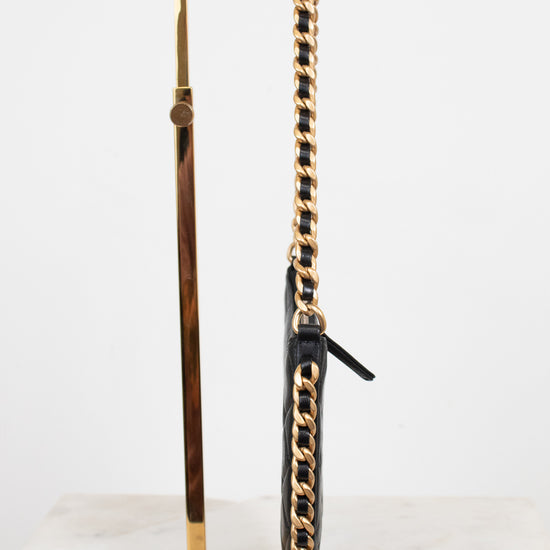 A black leather shoulder bag with a prominent gold chain strap, displayed on a gold metal stand against a white background.