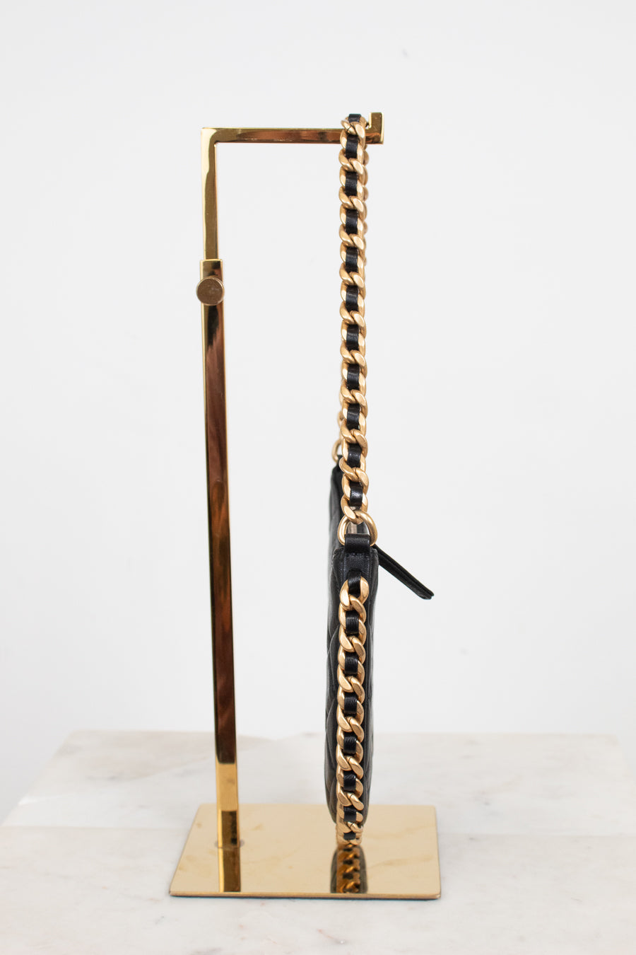A black leather shoulder bag with a prominent gold chain strap, displayed on a gold metal stand against a white background.