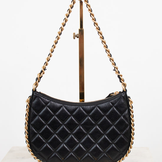 A black quilted leather shoulder bag with gold-tone chain straps, displayed on a gold metal stand against a white background.