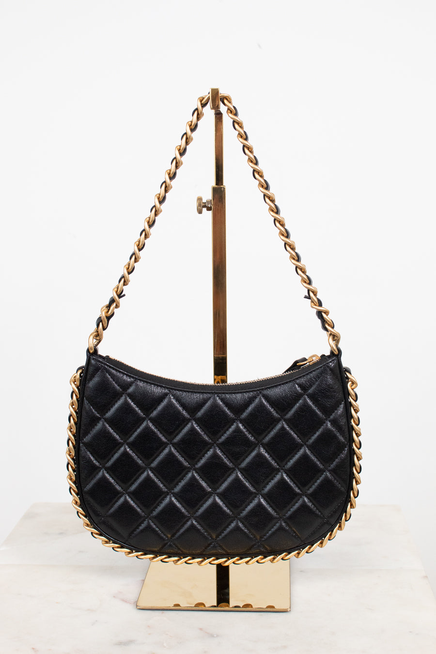 A black quilted leather shoulder bag with gold-tone chain straps, displayed on a gold metal stand against a white background.