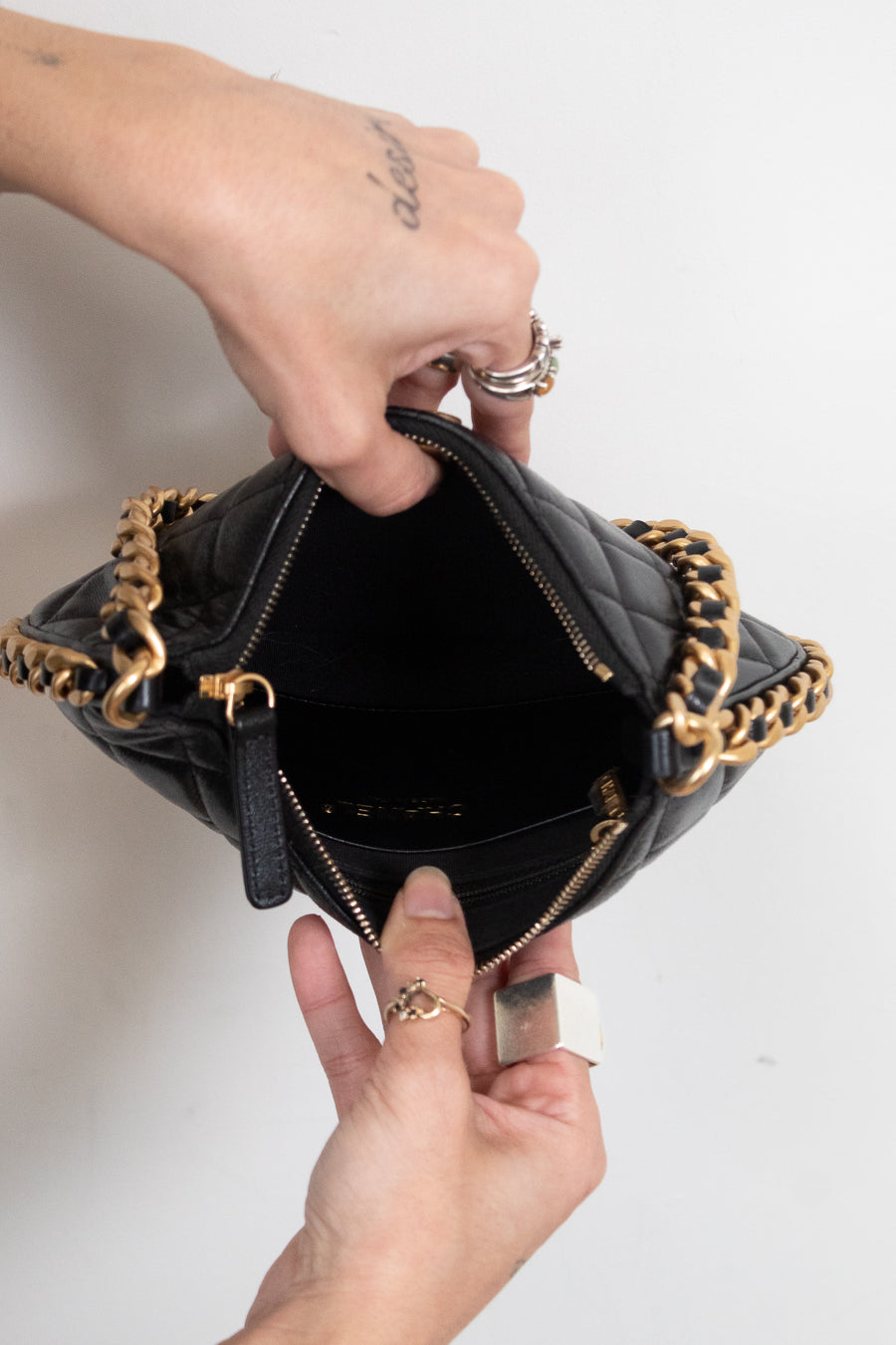 A close-up of a black quilted leather handbag with gold-tone chain straps, held open by two hands against a plain white background.