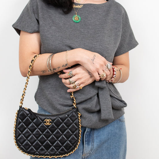 A woman wearing a gray t-shirt and blue jeans carries a black quilted Chanel shoulder bag with gold hardware and a gold chain strap.