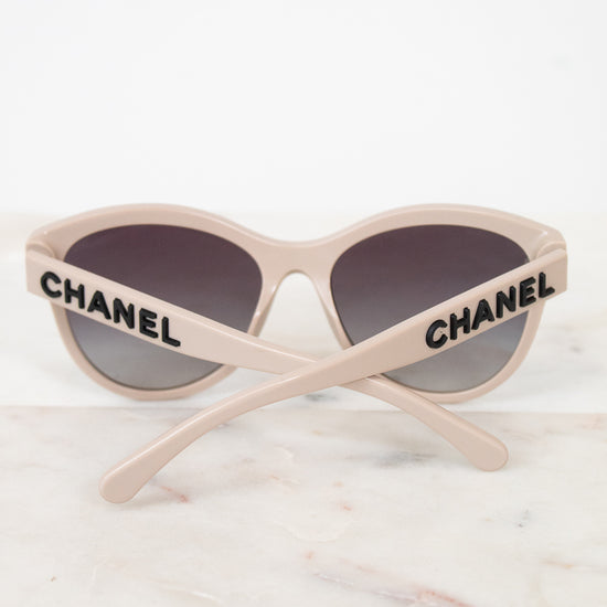 Chanel cat-eye sunglasses with a light beige frame, dark gray lenses, and black CHANEL lettering on the arms, resting on a white marble surface.