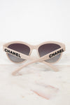 Chanel cat-eye sunglasses with a light beige frame, dark gray lenses, and black CHANEL lettering on the arms, resting on a white marble surface.