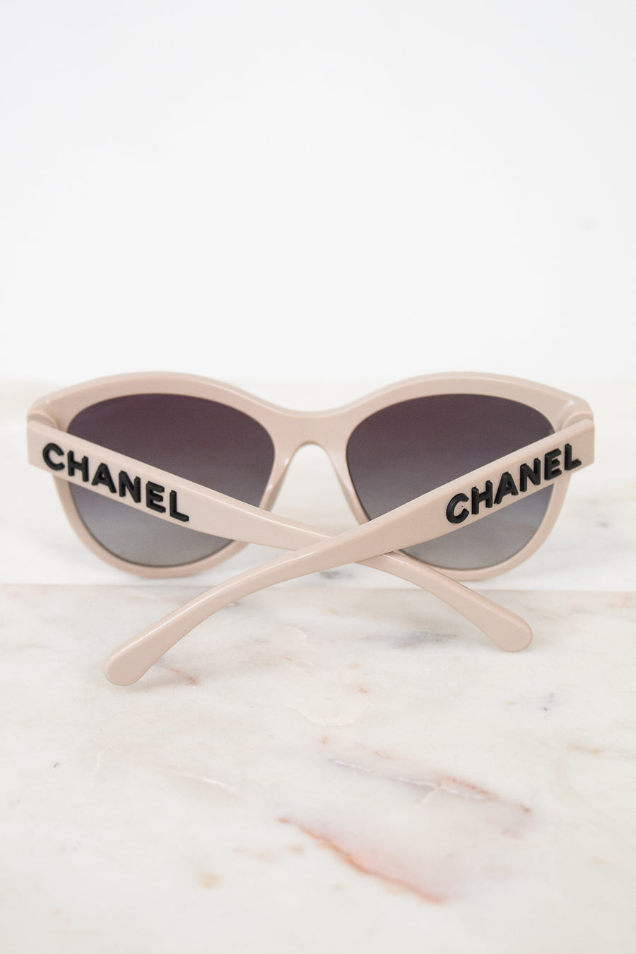 Chanel cat-eye sunglasses with a light beige frame, dark gray lenses, and black CHANEL lettering on the arms, resting on a white marble surface.
