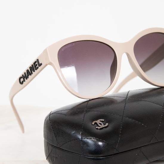 Chanel sunglasses with a beige frame and gradient gray lenses, resting on a black quilted leather case with a silver interlocking CC logo.