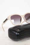 Chanel sunglasses with a beige frame and gradient gray lenses, resting on a black quilted leather case with a silver interlocking CC logo.