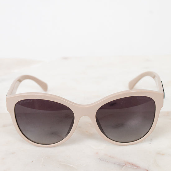 A pair of cat-eye sunglasses with a matte light beige frame and dark gray lenses, resting on a white marble surface against a white background.