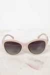 A pair of cat-eye sunglasses with a matte light beige frame and dark gray lenses, resting on a white marble surface against a white background.