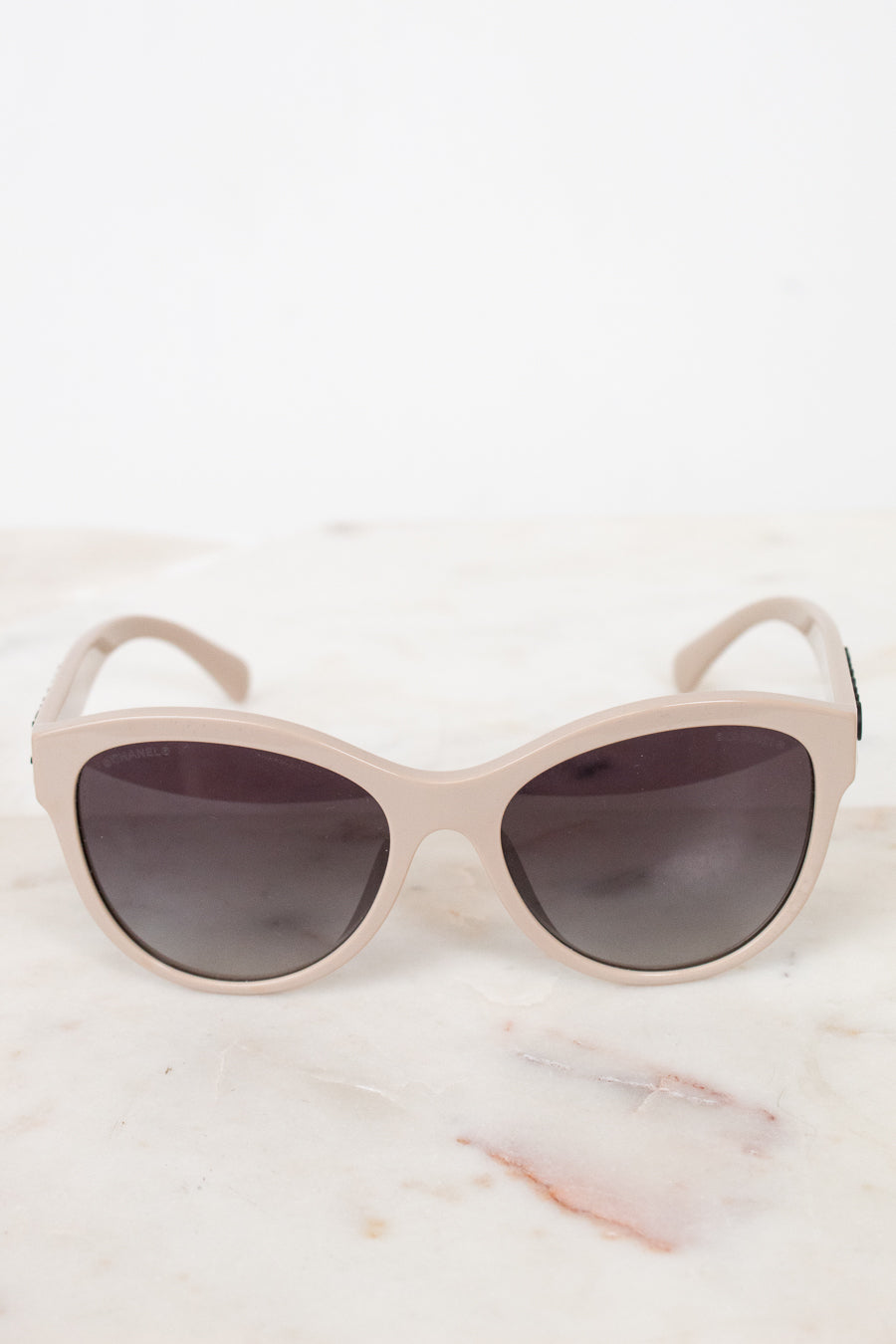A pair of cat-eye sunglasses with a matte light beige frame and dark gray lenses, resting on a white marble surface against a white background.