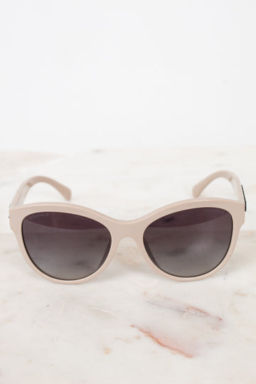 A pair of cat-eye sunglasses with a matte light beige frame and dark gray lenses, resting on a white marble surface against a white background.