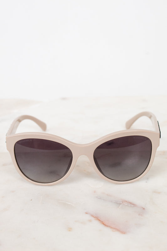A pair of cat-eye sunglasses with a matte light beige frame and dark gray lenses, resting on a white marble surface against a white background.
