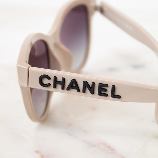 Close-up of Chanel sunglasses with a beige frame and dark lenses, resting on a white marble surface. The brand name CHANEL is printed in black on the side of the frame.