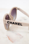 Close-up of Chanel sunglasses with a beige frame and dark lenses, resting on a white marble surface. The brand name CHANEL is printed in black on the side of the frame.