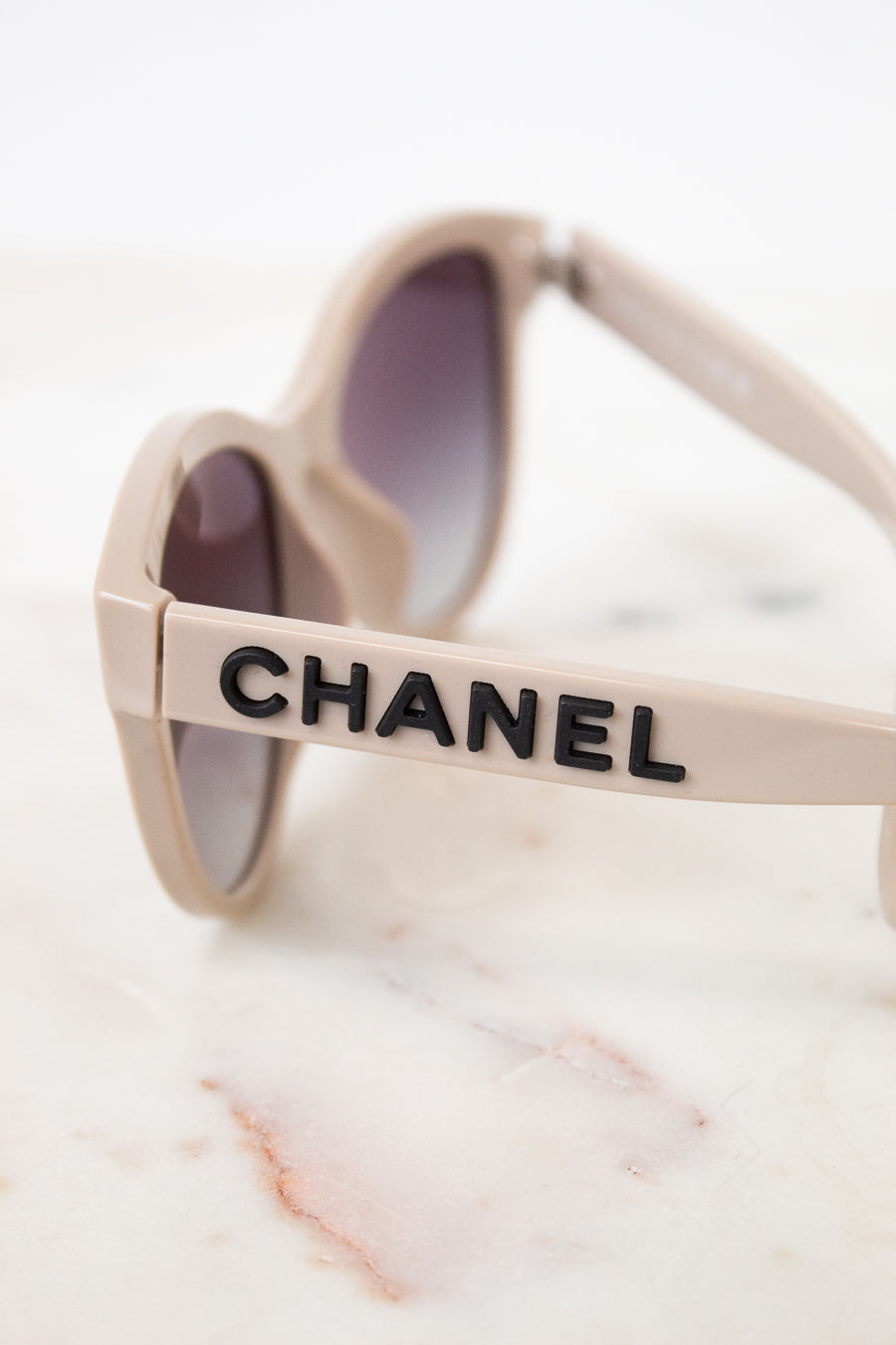 Close-up of Chanel sunglasses with a beige frame and dark lenses, resting on a white marble surface. The brand name CHANEL is printed in black on the side of the frame.
