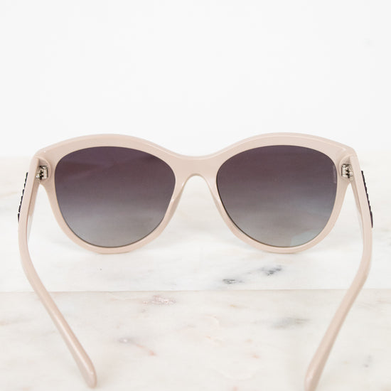 A pair of cat-eye sunglasses with a light beige frame and dark gradient lenses, displayed on a white marble surface against a white background.
