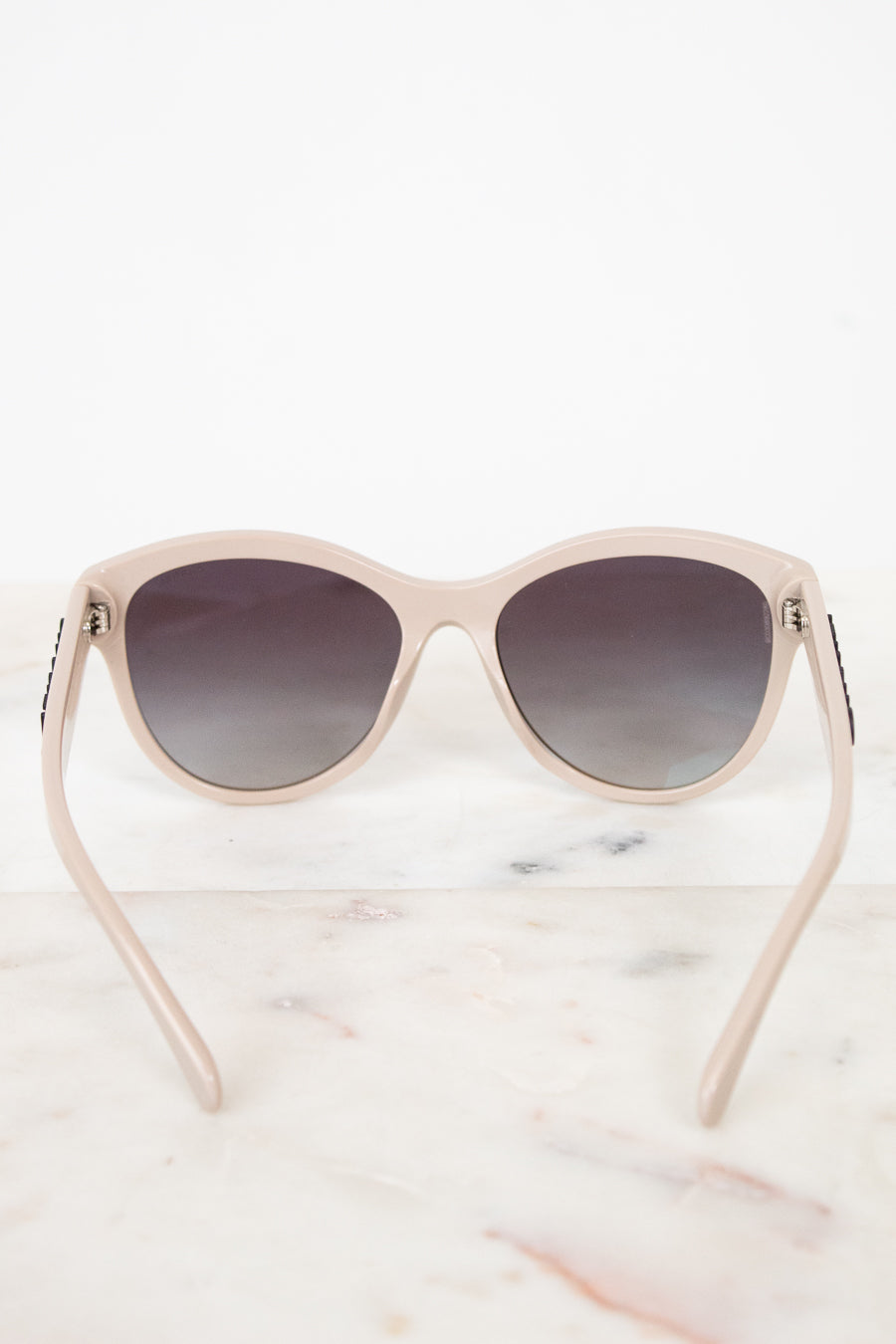 A pair of cat-eye sunglasses with a light beige frame and dark gradient lenses, displayed on a white marble surface against a white background.