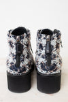 A pair of Chanel ankle boots with a black, white, and gray tweed pattern, featuring black platform soles and side zippers with the iconic interlocking CC logo.