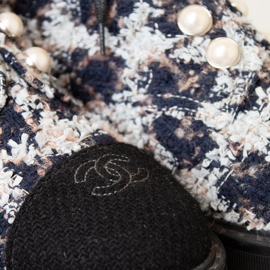 Close-up of a Chanel ankle boot featuring a navy, beige, and white tweed pattern, black laces, decorative pearl embellishments, and a black fabric cap with the iconic interlocking CC logo.
