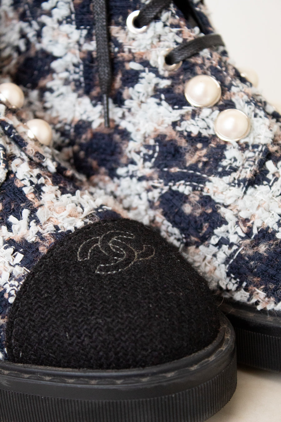 Close-up of a Chanel ankle boot featuring a navy, beige, and white tweed pattern, black laces, decorative pearl embellishments, and a black fabric cap with the iconic interlocking CC logo.
