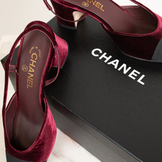 A pair of Chanel slingback high heels in deep burgundy velvet with black pointed toes, displayed on a black Chanel shoebox over a light marble surface.