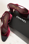 A pair of Chanel slingback high heels in deep burgundy velvet with black pointed toes, displayed on a black Chanel shoebox over a light marble surface.