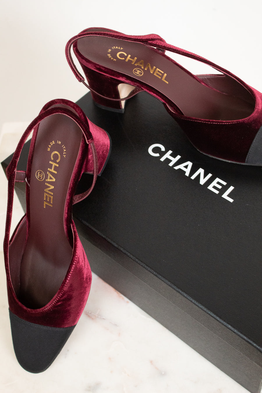 A pair of Chanel slingback high heels in deep burgundy velvet with black pointed toes, displayed on a black Chanel shoebox over a light marble surface.
